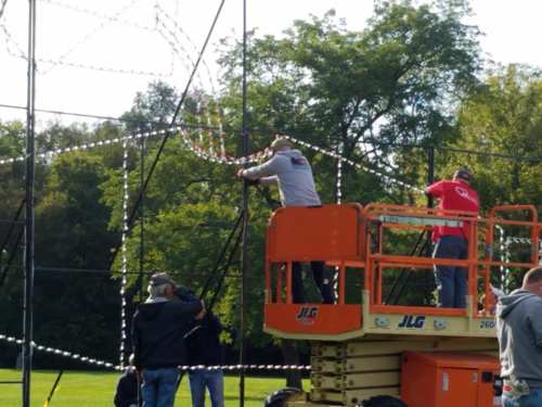 Workers on a lift install lights on a large structure in a park, surrounded by trees.