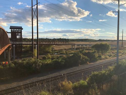 A scenic view of a railway track with a bridge, surrounded by greenery and under a blue sky with scattered clouds.