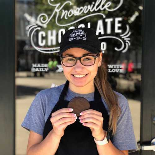 A smiling person in an apron holds a chocolate cookie in front of a chocolate shop window.