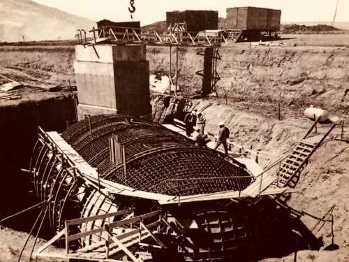 Black and white photo of construction workers on a large concrete structure with rebar, surrounded by earth and machinery.