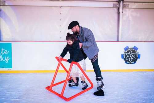 A man assists a young girl on the ice while she uses a skating aid for support.