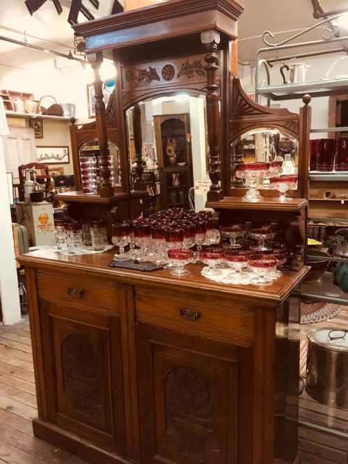 A vintage wooden sideboard with ornate mirrors and glassware displayed on top, set in an antique shop.