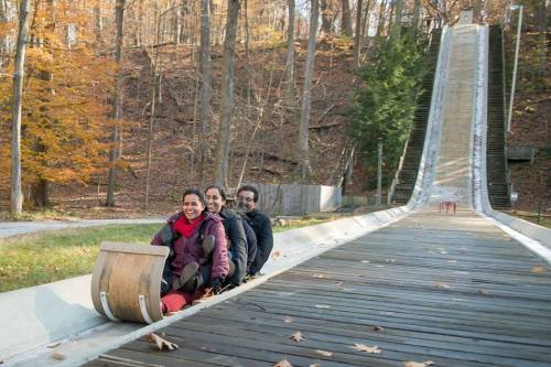 Three people sit on a sled at the base of a wooden toboggan run surrounded by autumn trees.