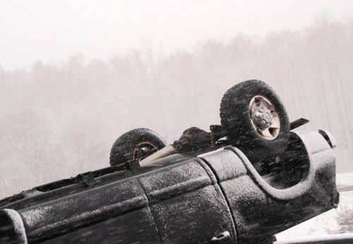 A black SUV overturned in a snowy landscape, with trees blurred in the background due to falling snow.