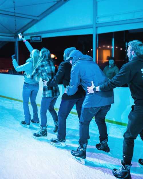 A group of people ice skating in a rink, holding onto each other while enjoying the activity under colorful lights.