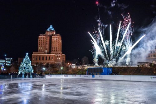 A festive ice rink at night with a lit Christmas tree and fireworks in the background, showcasing a winter celebration.