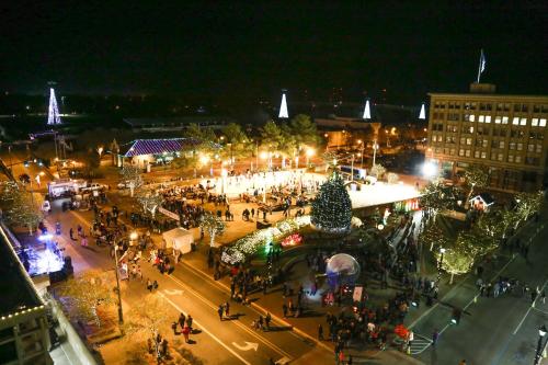 Aerial view of a festive outdoor ice skating rink at night, surrounded by holiday lights and a large crowd.