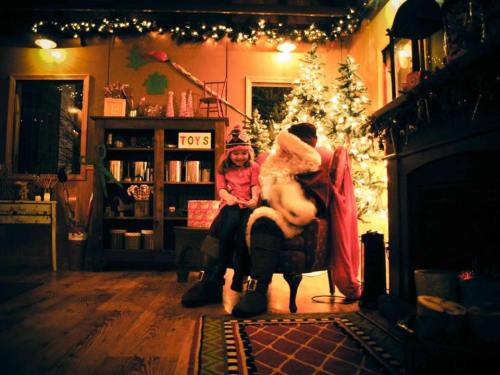 A cozy room with a Christmas tree, a child sitting on Santa's lap, and festive decorations in warm lighting.