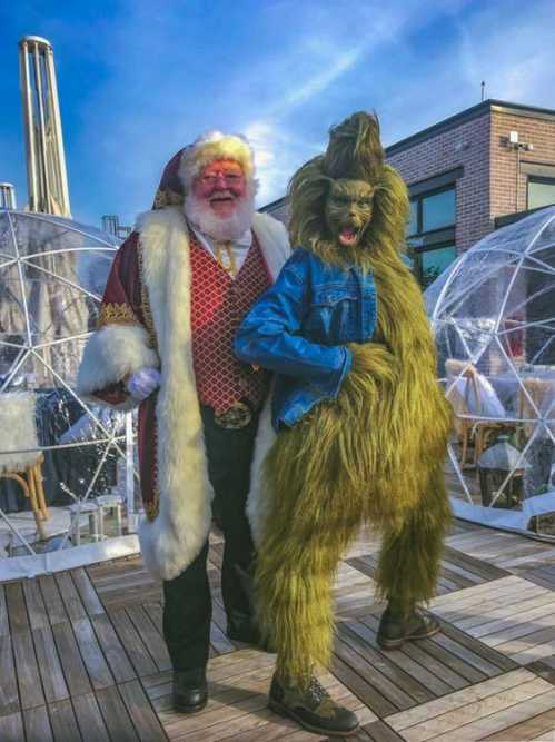 Santa Claus poses with a whimsical, furry creature in a denim jacket, set against a backdrop of clear igloos.