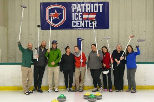 A group of nine people holding curling brooms and stones, smiling in front of a sign for the Patriot Ice Center.