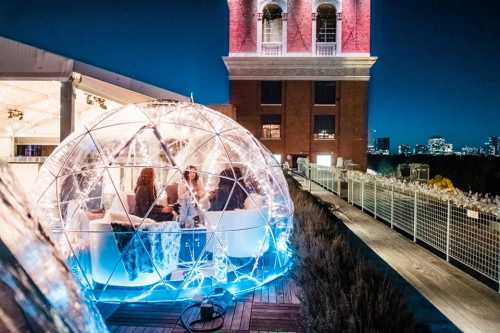 A cozy, illuminated igloo structure on a rooftop at night, with people dining and a city skyline in the background.