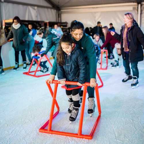 A mother helps her daughter skate on ice using a support frame, surrounded by other skaters in a festive indoor rink.