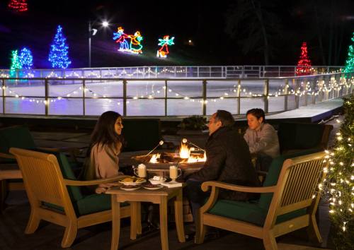 A cozy outdoor scene with three people around a fire pit, festive lights, and an ice skating rink in the background.