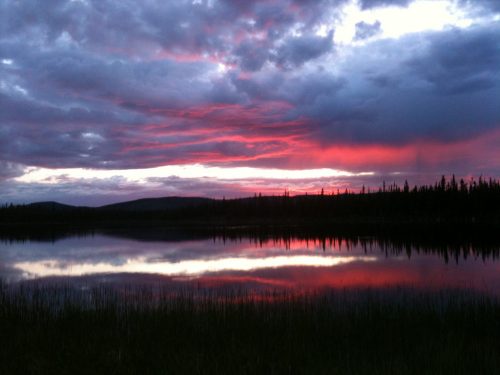 A serene lake at sunset, reflecting vibrant pink and purple clouds, surrounded by dark silhouettes of trees and hills.