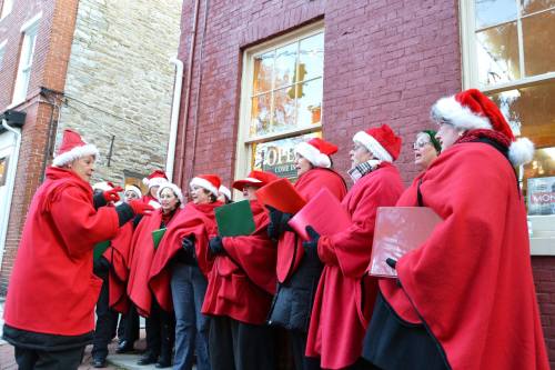 A group of carolers in red capes and Santa hats singing outside a brick building during the holiday season.