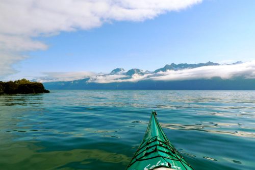 A kayak glides over calm waters, with mountains and clouds in the background under a clear blue sky.