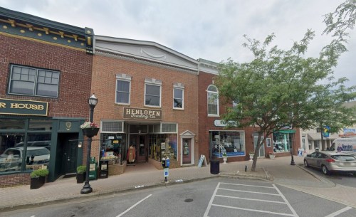 A brick storefront with "Henlopen Trading Co" sign, surrounded by trees and other shops on a cloudy day.