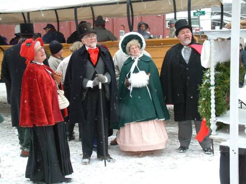 A group of four people in Victorian attire, smiling and standing in the snow, with festive decorations around them.