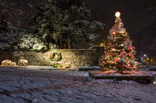 A snowy night scene featuring a decorated Christmas tree and a wreath, illuminated by festive lights.