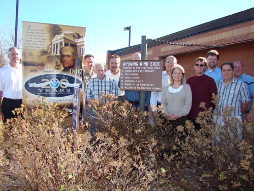 A group of people stands in front of a sign and banner celebrating 200 years, surrounded by shrubs.