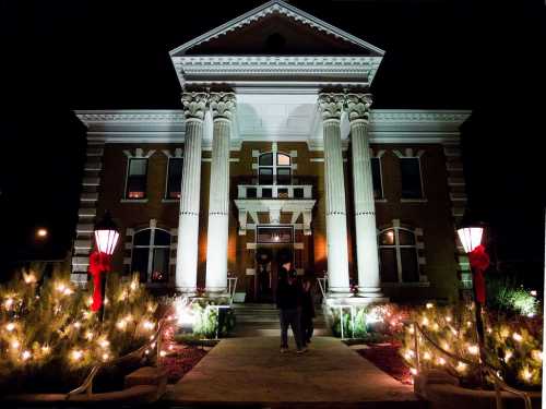 Historic building illuminated at night, adorned with festive lights and decorations, with two people walking in front.