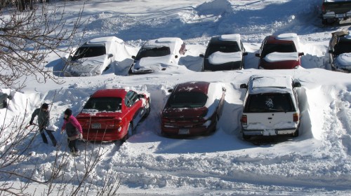 Two people shovel snow around parked cars in a snowy parking lot, with several vehicles partially buried in snow.