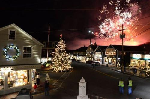 A festive town square at night, adorned with holiday lights and a Christmas tree, with fireworks lighting up the sky.