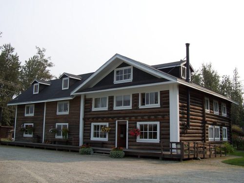 A large log cabin-style house with multiple sections, white trim, and flower boxes, set in a natural landscape.