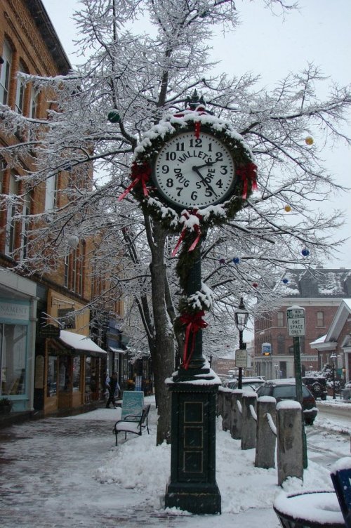 A snow-covered street scene featuring a decorative clock surrounded by festive decorations and trees.