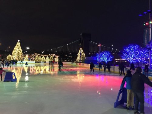 A festive ice skating rink at night, adorned with colorful lights and decorated trees, with a bridge in the background.