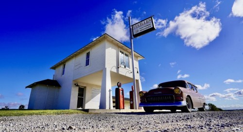 A vintage car parked in front of a classic gas station under a bright blue sky with fluffy clouds.