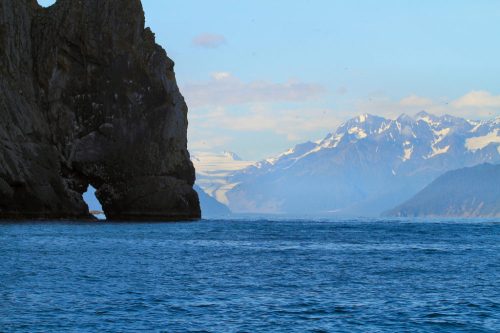 A rocky arch stands by the water, with snow-capped mountains and a clear blue sky in the background.