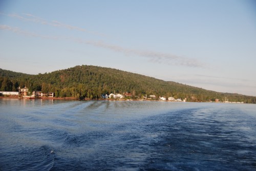 A serene lake scene with a forested hill and a small town along the shore, reflecting calm waters under a clear sky.