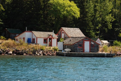 Quaint cabins with red trim by a calm lake, surrounded by lush green trees and rocky shoreline.