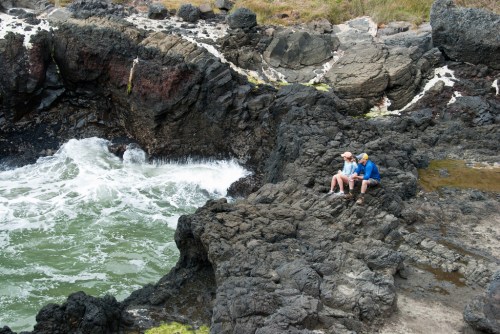 Two people sit on rocky shore, overlooking waves crashing against the rocks in a coastal landscape.