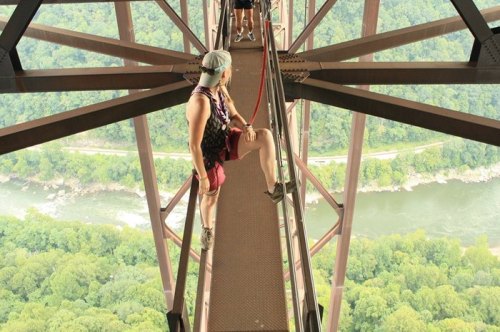 A person stands on a narrow metal walkway high above a river, surrounded by lush green trees and a scenic landscape.