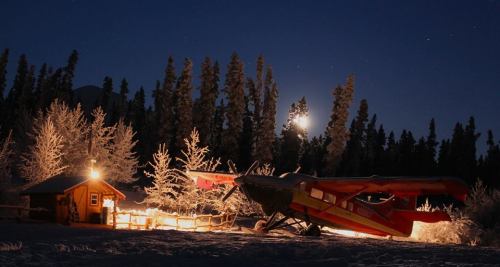 A colorful plane parked near a cozy cabin, surrounded by snow-covered trees under a starry night sky.