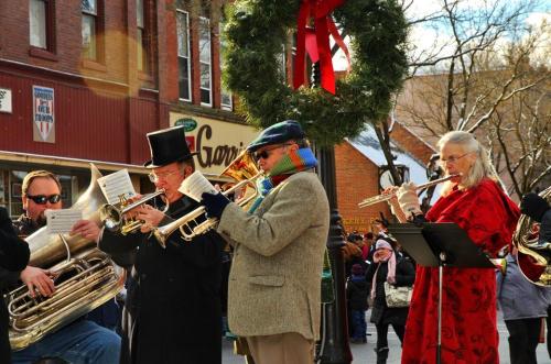 A festive street scene with musicians playing brass instruments, wearing winter attire, and surrounded by holiday decorations.