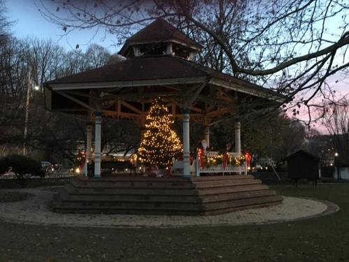 A decorated gazebo with a Christmas tree, surrounded by trees, at dusk with festive lights.