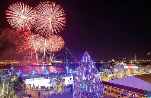 Colorful fireworks illuminate the night sky over a festive waterfront scene with holiday lights and an ice skating rink.