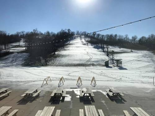 A snowy ski slope under a clear blue sky, with empty benches in the foreground and ski lifts in the distance.