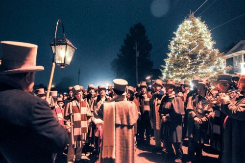 A group of carolers in top hats and scarves sings near a brightly lit Christmas tree at night.