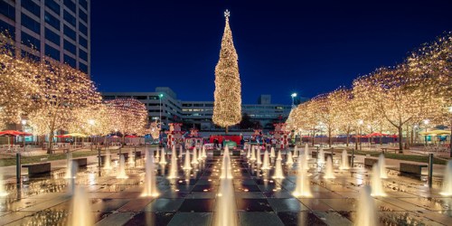 A festive plaza at night, featuring a tall, illuminated Christmas tree and decorated snowmen, surrounded by glowing lights.