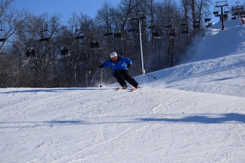 A skier in a blue jacket glides down a snowy slope with ski lifts in the background on a clear day.