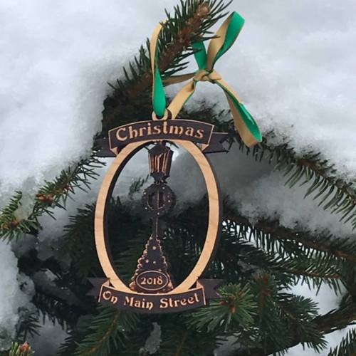 A wooden Christmas ornament reading "Christmas on Main Street 2018," hanging from a snowy evergreen branch.