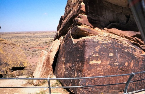 Petroglyphs etched into a large rock formation, surrounded by a vast desert landscape under a clear blue sky.