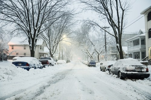 A snowy street lined with houses and parked cars, blanketed in fresh snow during a winter storm.