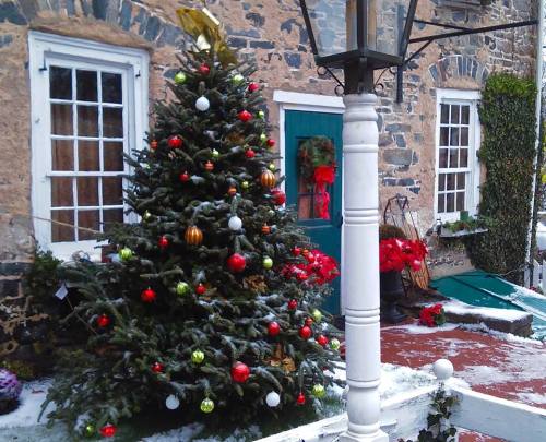 A decorated Christmas tree stands outside a stone building, surrounded by snow and festive decorations.
