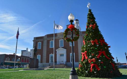 A festive Christmas tree adorned with decorations stands in front of a brick building, with flags and a statue nearby.