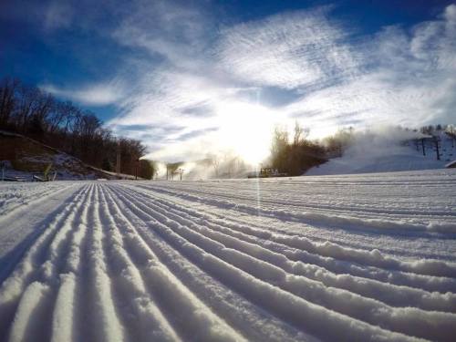 A snowy landscape with groomed ski trails under a bright sky and sun, surrounded by trees.